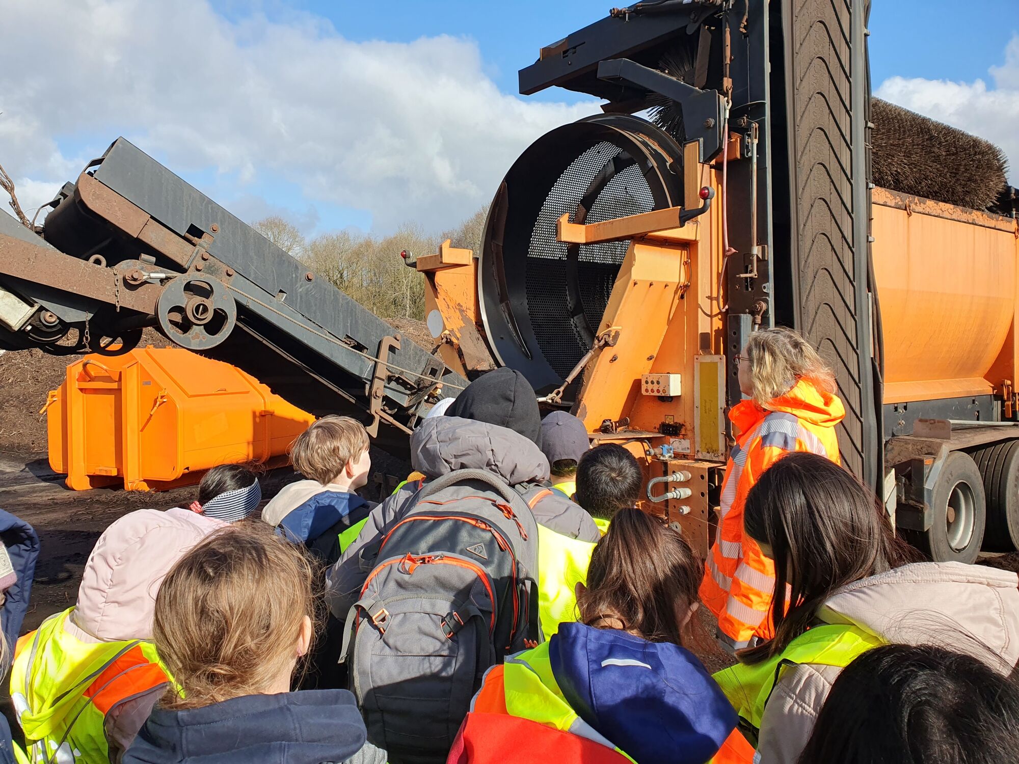  – Eine Frau in orangener Sicherheitsjacke und Kinder, die auf ein orangefarbenes Trommelsieb gucken.
