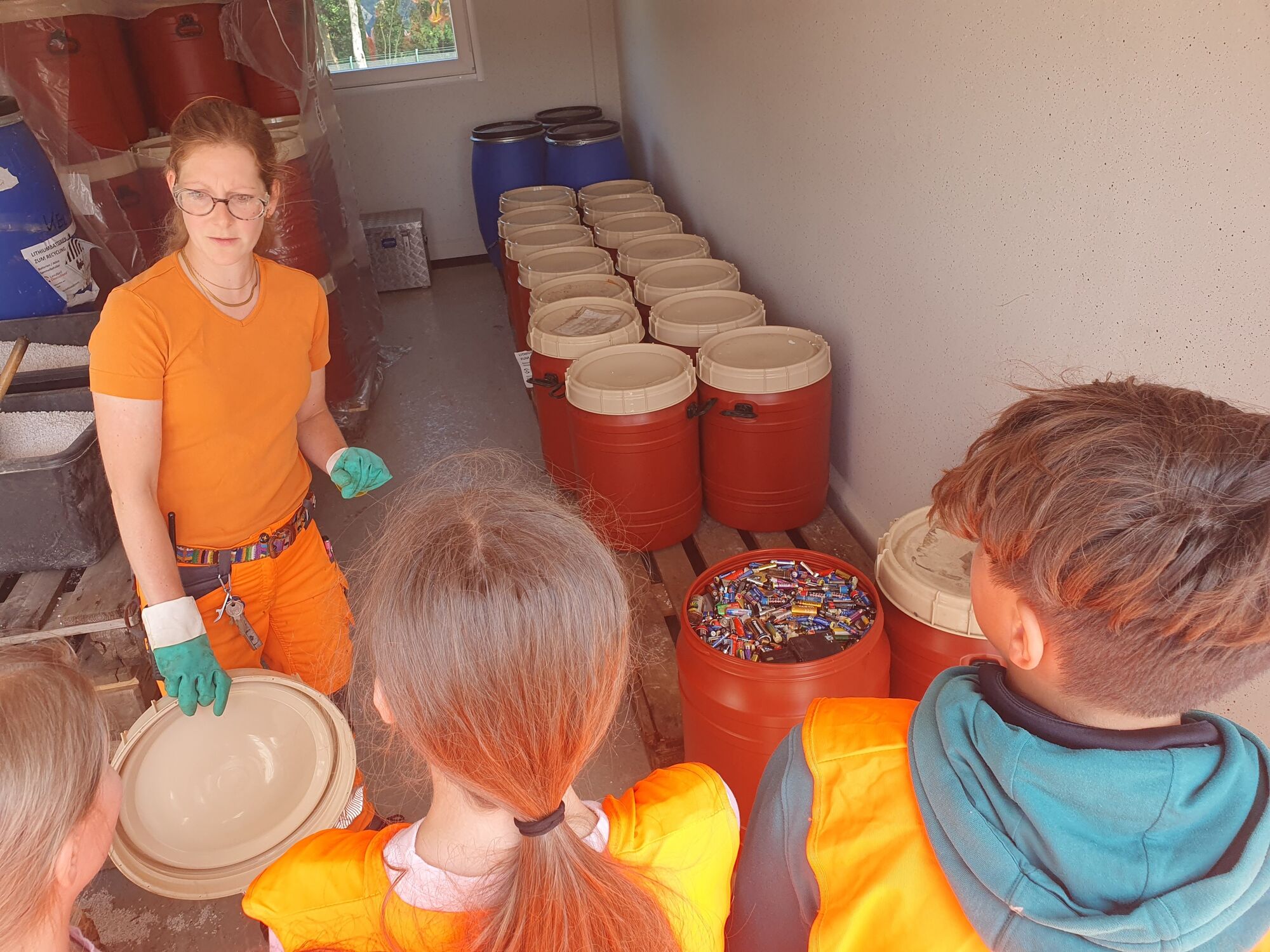  – Eine Frau in orangener Arbeitskleidung und grünen Arbeitshandschuhen steht in einem Container und zeigt Kindern etwas. Sie hält einen Deckel von einem Fass in der Hand. Neben ihr  steht ein geöffnetes rotes Fass mit kleinen Batterien. Hinter ihr in dem Container sind rote und blaue Spannringdeckelfässer.