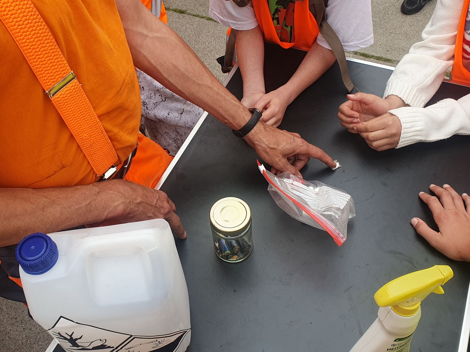  – Auf einem Tisch stehen ein Plastikkanister, ein kleines Glas mit Batterien, eine Plastiktüte und eine Knopfzelle, um die durchsichtiges Klebeband gewickelt ist. Die Hand einer Person in orangefarbener Arbeitskleidung zeigt auf diese Knopfzelle.