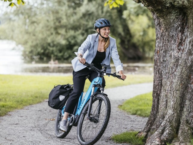 Radfahrerin fährt mit einem E-Bike durch einen Park. Sie hat einen Helm auf und Packtaschen am Rad.