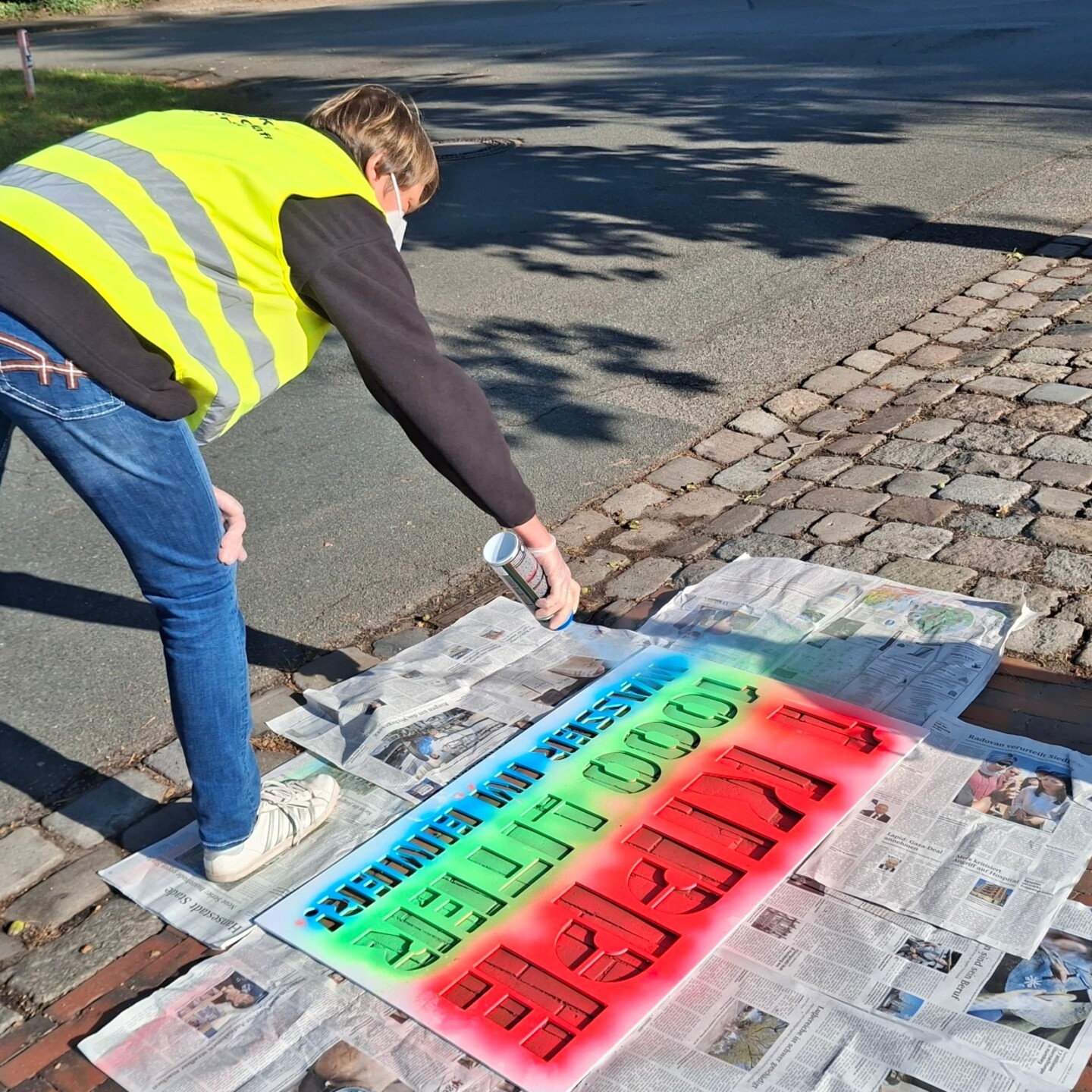 Eine Person mit neonfarbener Sicherheitsweste spr&uuml;ht einen Spruch in den Farben rot, gr&uuml;n und blau mittels einer Schablone einen Gehweg neben einer Strasse.