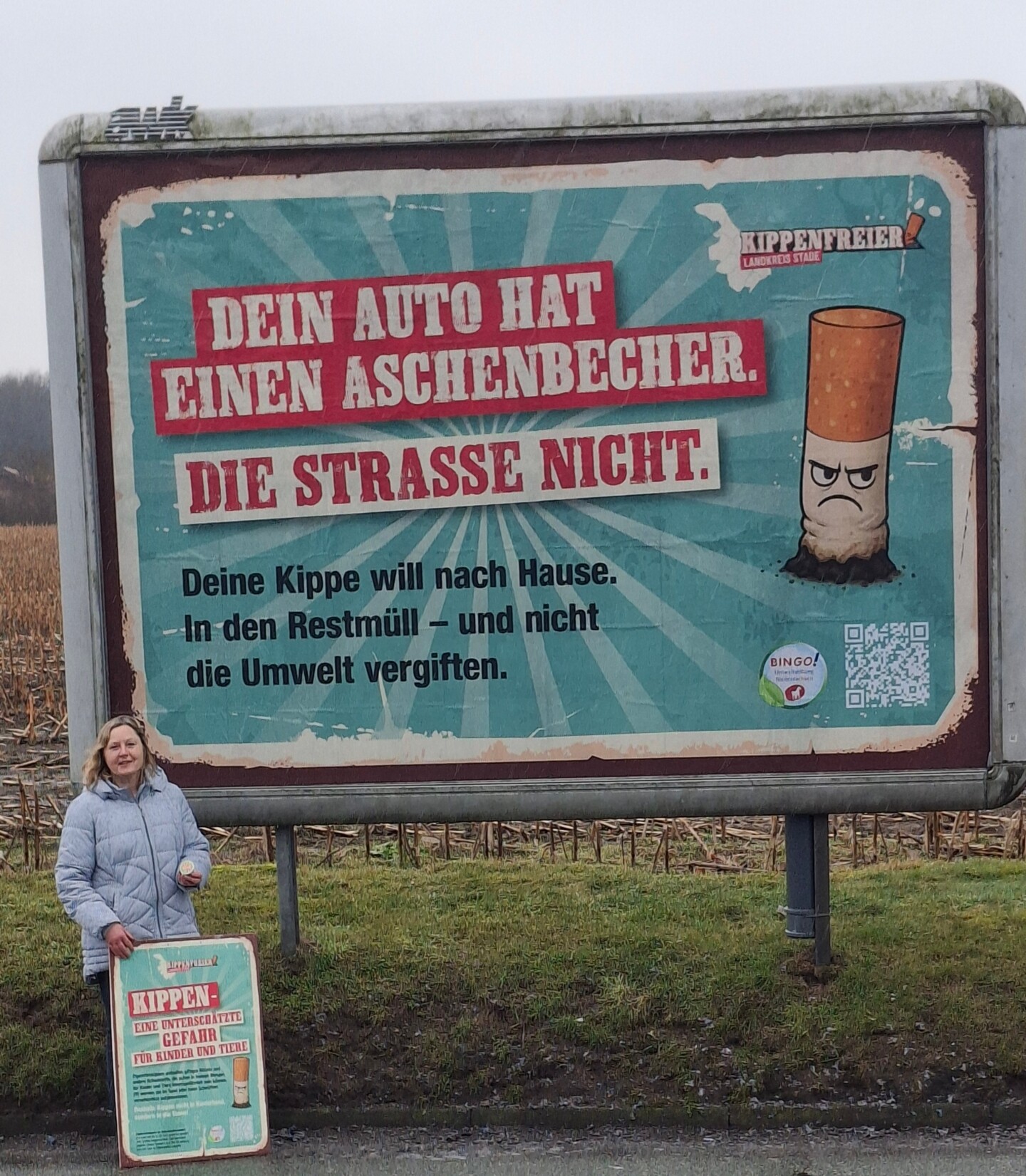 Eine Frau, die ein Plakat vor sich hält, steht vor einem Großplakat. Darauf steht in großer Schrift auf rotem Hintergrund 