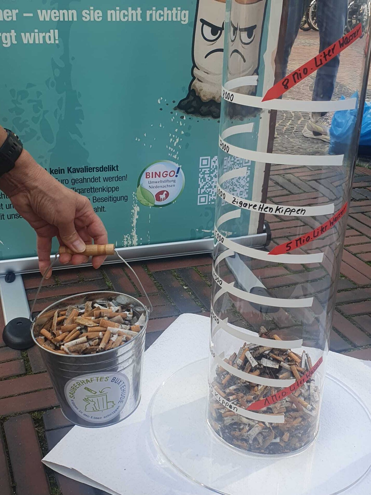 Eine Plexiglass&auml;ule mit einigen Zigarettenkippen sowie ein kleiner Metalleimer, der mit Zigarettenkippen gef&uuml;llt ist.