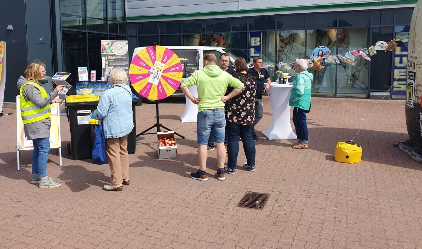 Ein Informationsstand mit zwei Stehtischen, Glücksrad, Kundenstopper, Mülltonne. Mehrere Personen stehen davor und unterhalten sich.