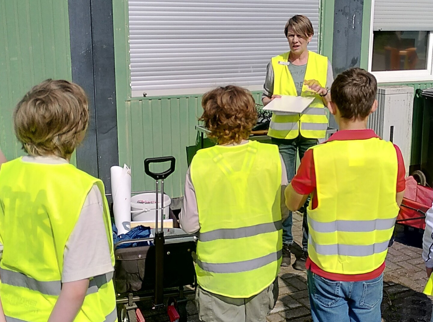 Eine Frau, die eine Sicherheitsweste trägt, neben ihr ein Bollerwagen mit Materialien, erzählt drei Kindern, die auch Sicherheitswesten tragen, etwas. Sie stehen vor einem grünlichen Container.