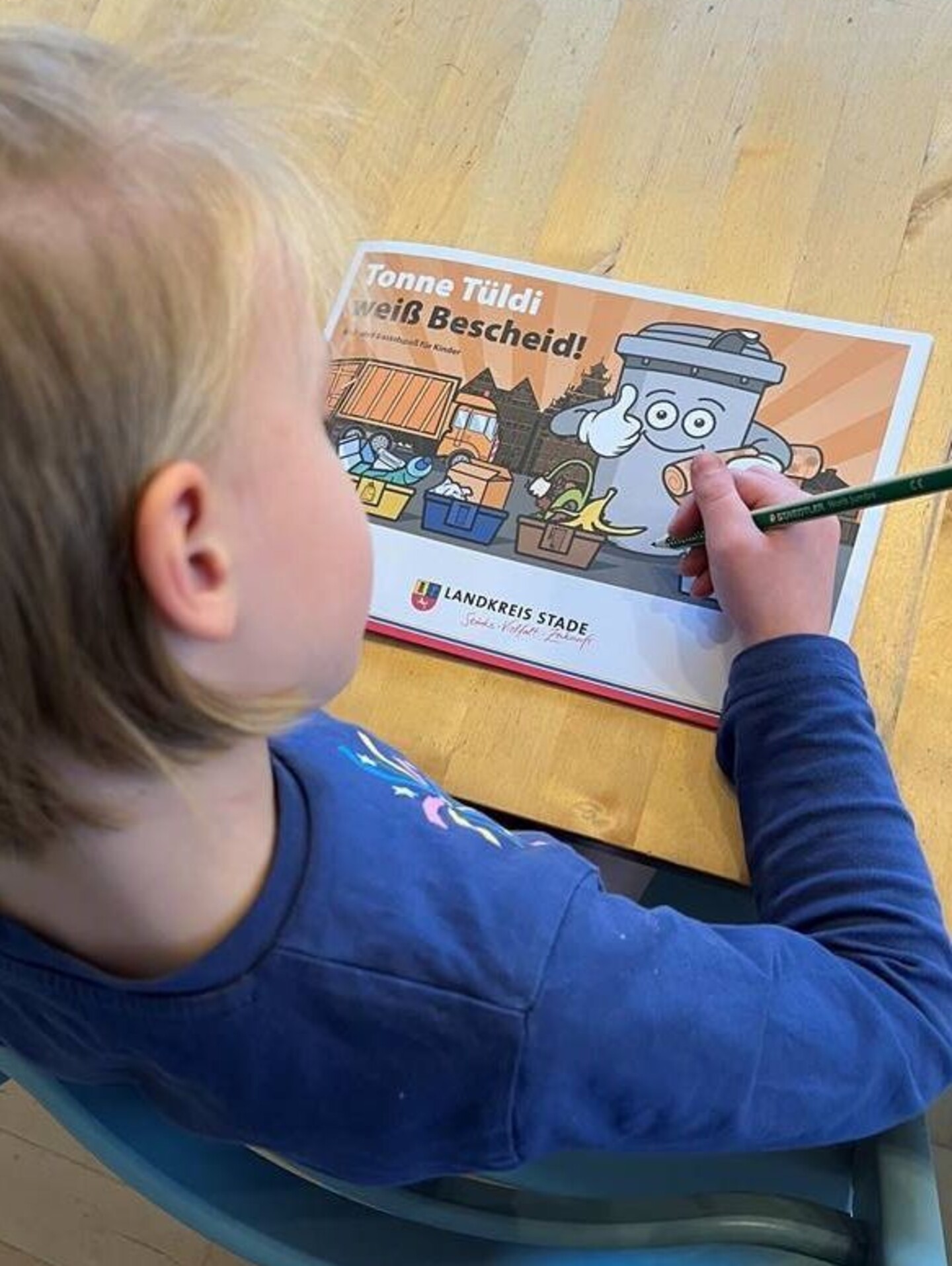 Ein Kind mit einem Bleistift in der Hand- Vor ihr auf einem Tisch das Mal- und Bastelheft Tonne Tüldi weiß Bescheid des Landkreises Stade.