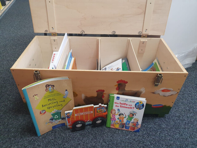 Eine aufgeklappte Holzkiste mit verschiedenen Fächern. In den Fächern&nbsp; und vor der Kiste liegen Kinderbücher.