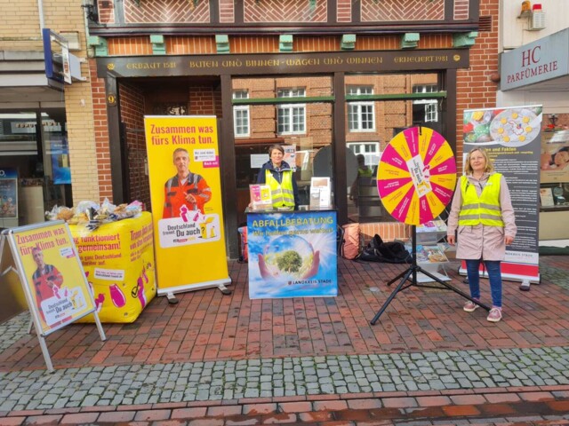 Informationsstand mit Roll-up, Glückrad, Informationstresen, aufgeblasenem gelben Würfel und Kundenstopper. Eine Frau steht hinter dem Tresen, die andere vor einem Roll up.&nbsp;