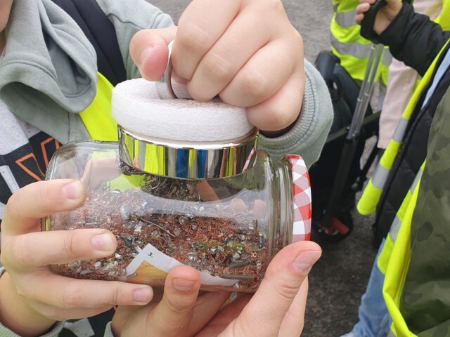 Kinderhände halten ein Glas, in dem silbrige und rötliche Metallspäne ist, in den Händen. Daran halten sie einen Magneten.