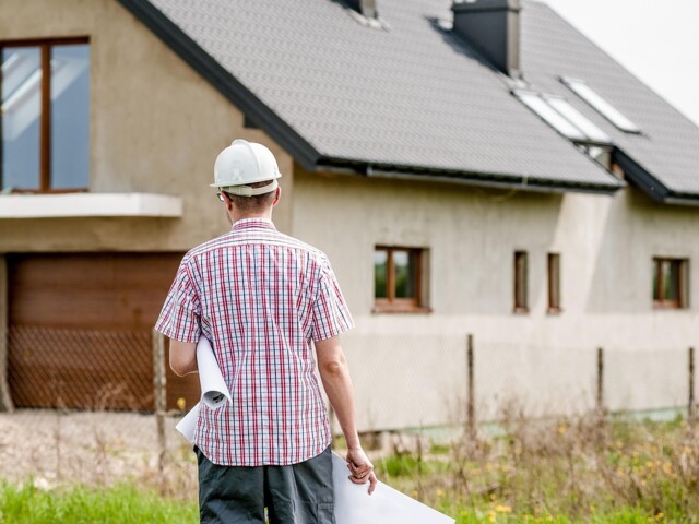 Mann mit Helm und Papieren in der Hand steht vor einem Haus